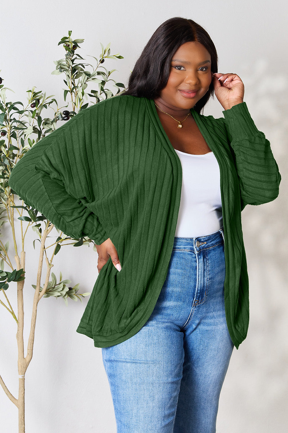 A woman confidently poses in a Basic Bae Full Size Ribbed Cocoon Cardigan in green, crafted from slightly stretchy polyester, over a white top and paired with blue jeans. She stands beside a leafy plant, gently touching her hair and smiling warmly at the camera, perfectly embodying a casual yet basic style.