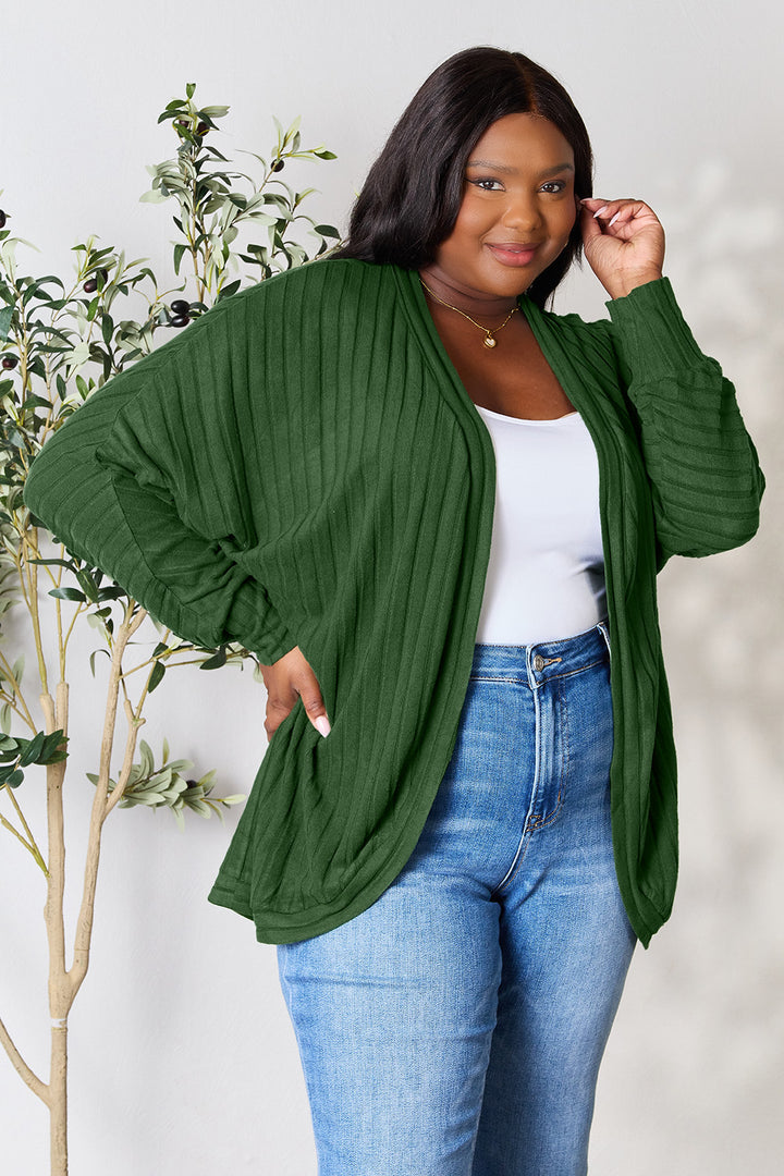 A woman confidently poses in a Basic Bae Full Size Ribbed Cocoon Cardigan in green, crafted from slightly stretchy polyester, over a white top and paired with blue jeans. She stands beside a leafy plant, gently touching her hair and smiling warmly at the camera, perfectly embodying a casual yet basic style.