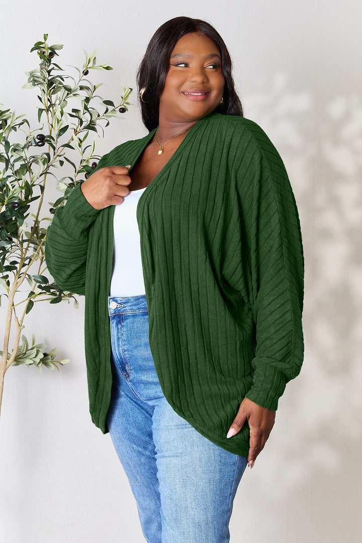 A person wearing the Basic Bae Full Size Ribbed Cocoon Cardigan in green over a white top and blue jeans stands smiling near a plant against a light-colored wall. The slightly stretchy cardigan, made of polyester, drapes stylishly, and the person embraces basic style with their long dark hair flowing freely.