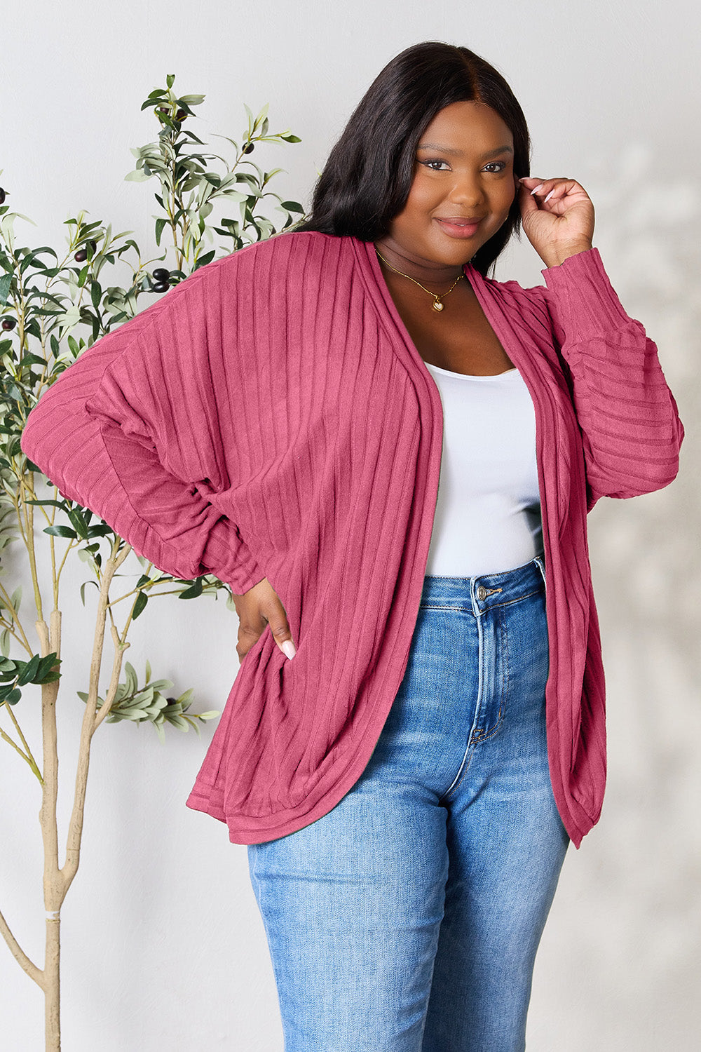 Dressed in the Basic Bae Full Size Ribbed Cocoon Cardigan, a person confidently stands against a light grey background adorned with a leafy plant. The pink cardigan, made from slightly stretchy polyester, is stylishly paired with a white top and blue jeans. Embracing a basic style, they smile warmly while touching their hair.