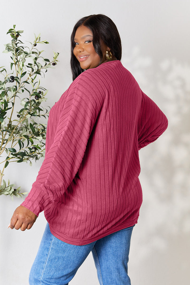 A person with long dark hair is wearing a slightly stretchy, ribbed red cardigan from the Basic Bae Full Size Ribbed Cocoon collection and blue jeans. They are standing sideways, smiling in front of a light-colored background with a potted plant.