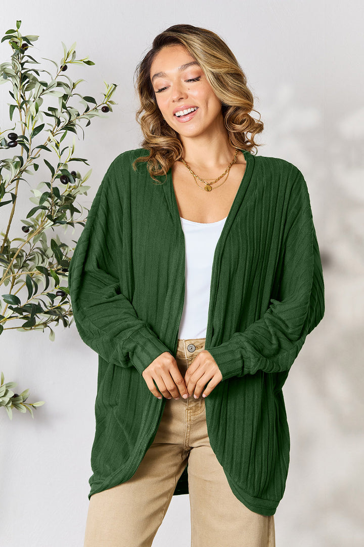 A person with wavy hair smiles while wearing a slightly stretchy Basic Bae Full Size Ribbed Cocoon Cardigan in green, layered over a white top and beige pants. They stand in a basic style beside a leafy plant against a light background.