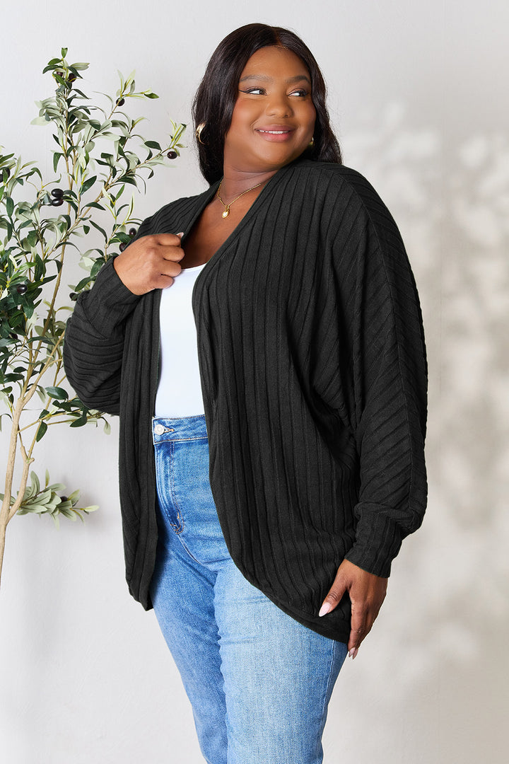 A woman smiles while wearing the Basic Bae Full Size Ribbed Cocoon Cardigan in black over a white top and blue jeans. She stands next to a potted plant against a light-colored wall, exuding an effortlessly chic basic style.