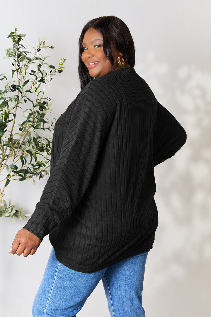 A person with long dark hair is smiling, wearing the Basic Bae Full Size Ribbed Cocoon Cardigan, made of slightly stretchy polyester, along with blue jeans. They are standing in front of a light-colored background with a plant on the left side, embracing a basic style.