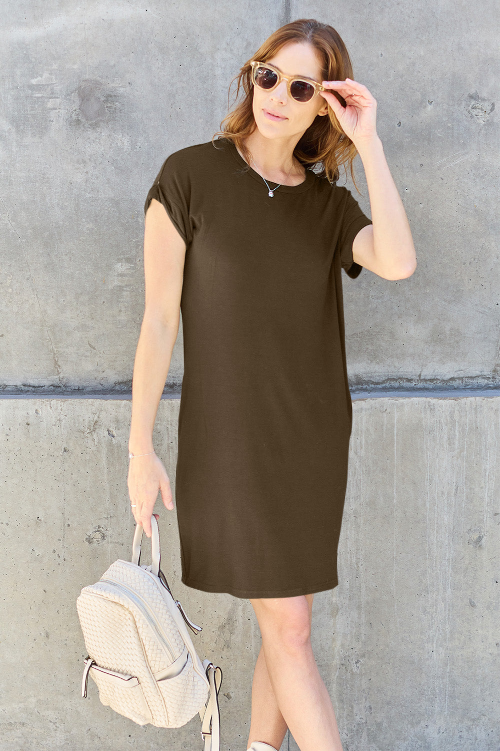 A person with long hair in sunglasses and a casual black Basic Bae Full Size Round Neck Short Sleeve Dress with Pockets leans against a concrete wall. They hold a textured polyester backpack in one hand and adjust their sunglasses with the other.