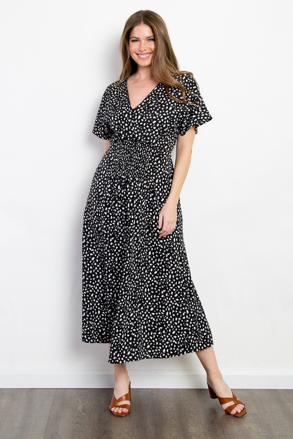 A woman in a Be Stage Full Size Printed Smocked Waist Dress smiles against a white wall. Shes wearing brown sandals with her long hair down. The knee-length dress features short sleeves and an eye-catching print, making it versatile for any occasion.