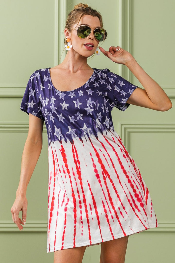 A woman confidently poses in front of a light green paneled wall, donning the BiBi American Flag Theme Tee Dress featuring a star-patterned blue top and red-striped white bottom, embodying the spirit of Americana fashion. Her hand gently rests on her sunglasses.