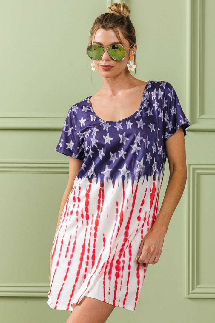 Dressed in a BiBi American Flag Theme Tee Dress, the individual showcases an Americana look with sunglasses, hoop earrings, and a bun against a light green wall—a starry blue top paired with red stripes on a white bottom.