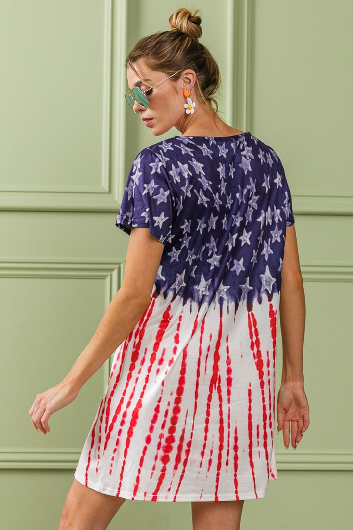 A woman in sunglasses poses in the BiBi American Flag Theme Tee Dress, with a navy top adorned with white stars transitioning into red and white stripes. Her hair is stylishly in a bun, standing against a light green paneled wall, embodying Americana fashion.