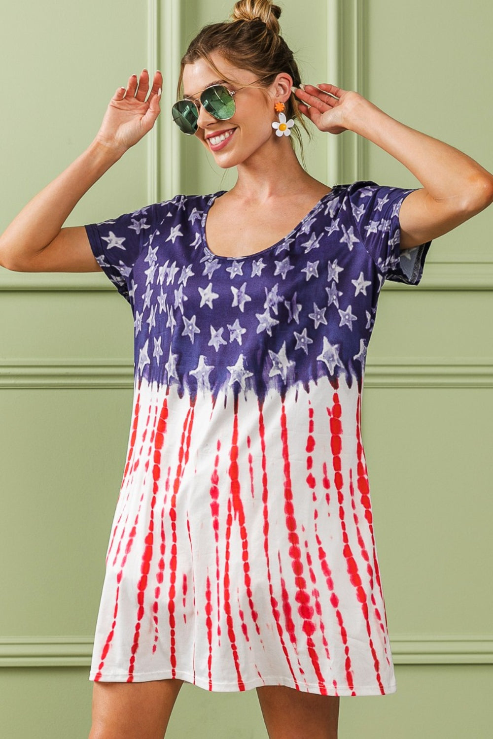 A woman in star earrings and sunglasses wears a BiBi American Flag Theme Tee Dress, featuring a blue backdrop with stars and red stripes. She smiles while playing with her hair against a light green paneled wall.