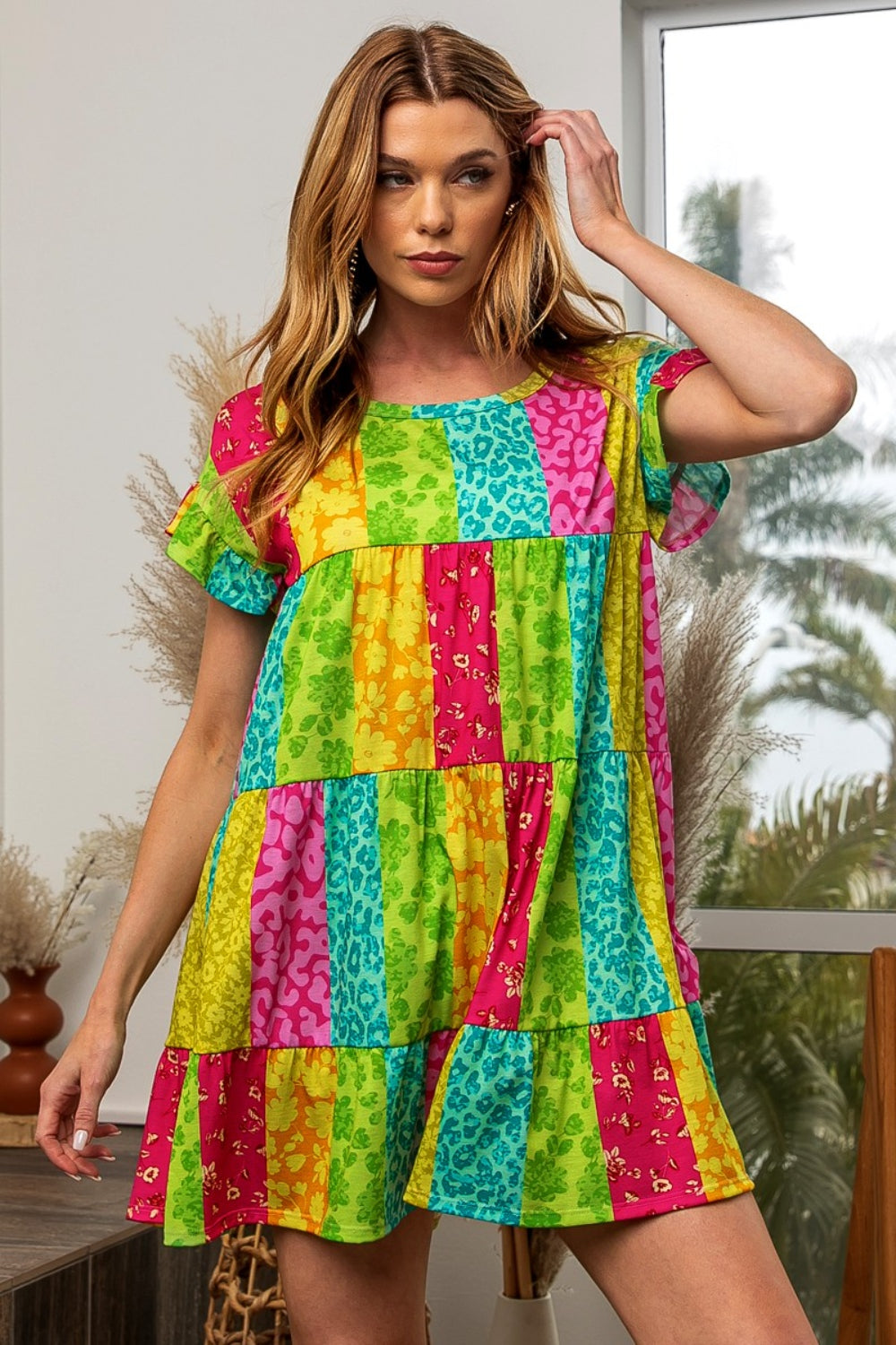 A person with long hair wears the BiBi Leopard Short Sleeve Tiered Dress indoors, featuring a colorful patchwork pattern of bright stripes in green, yellow, pink, and blue. Plants and a window with greenery are visible in the background.