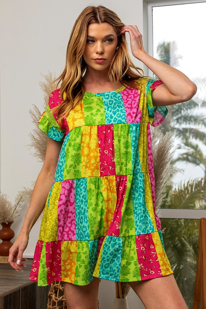 A person with long hair wears the BiBi Leopard Short Sleeve Tiered Dress indoors, featuring a colorful patchwork pattern of bright stripes in green, yellow, pink, and blue. Plants and a window with greenery are visible in the background.