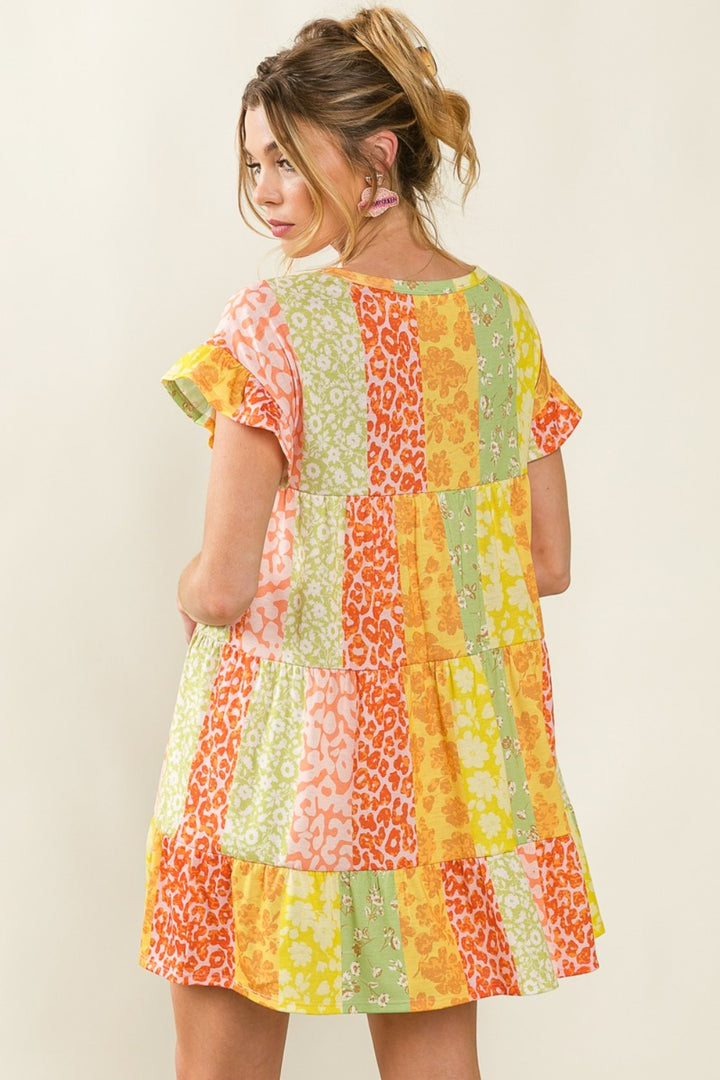 In a vibrant BiBi Leopard Short Sleeve Tiered Dress with colorful floral and animal prints, including bold leopard patches, a woman gazes over her shoulder. Her light brown hair in a ponytail has loose strands complementing her pink earrings and the dresss yellow, orange, pink, and green hues.