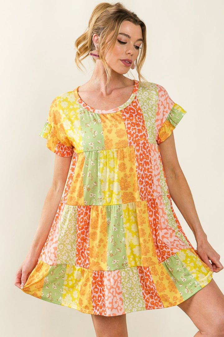 A woman models the BiBi Leopard Short Sleeve Tiered Dress, displaying its vibrant patchwork in green, orange, pink, and yellow. The dress features floral and leopard prints. With hair styled up, she holds the sides and looks down.
