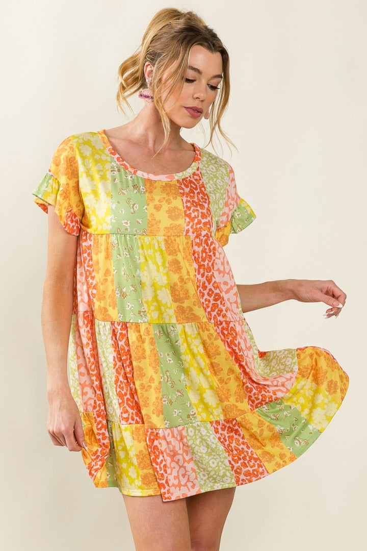 A woman in a BiBi Leopard Short Sleeve Tiered Dress, featuring vibrant orange, yellow, green, and pink floral and animal prints. She holds the hem, wearing her hair in a loose updo with pink earrings against a plain light background.