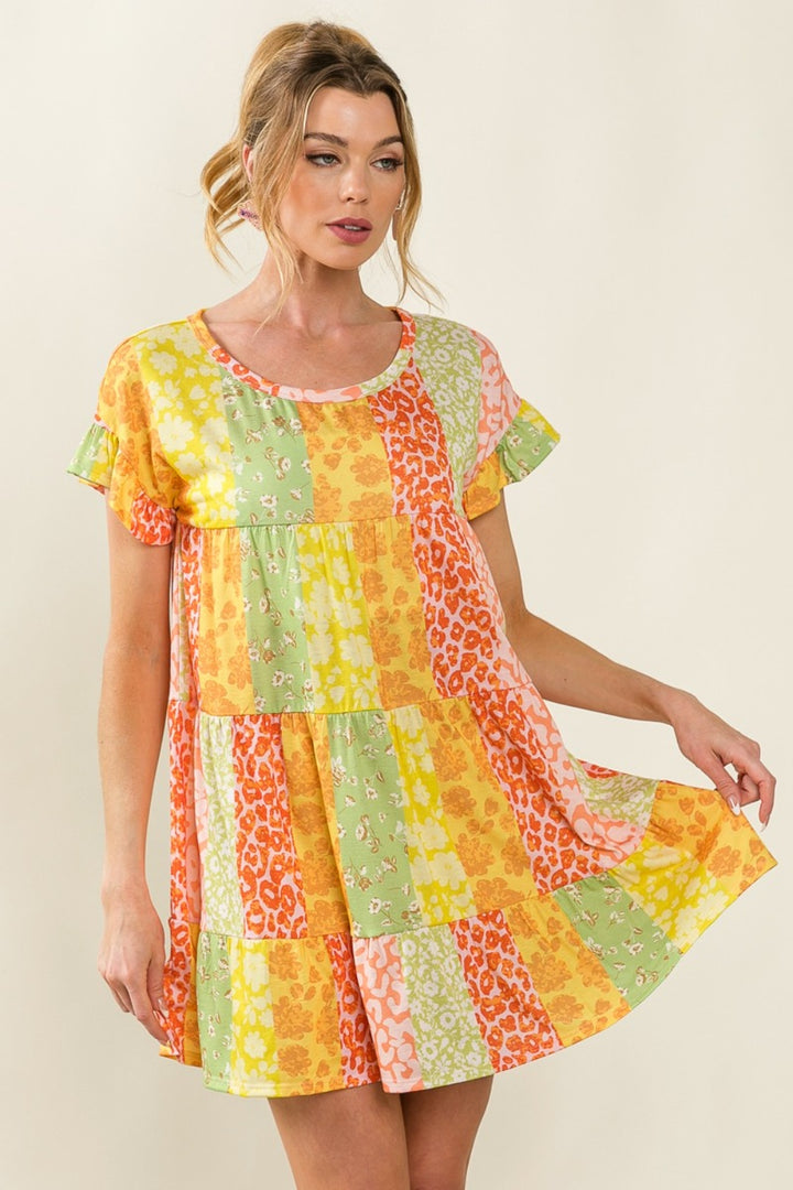 A woman models the BiBi Leopard Short Sleeve Tiered Dress, featuring a vibrant patchwork of orange, yellow, green, and pink. With floral and leopard prints, she gracefully holds the hem of her dress while sporting an elegant updo hairstyle.