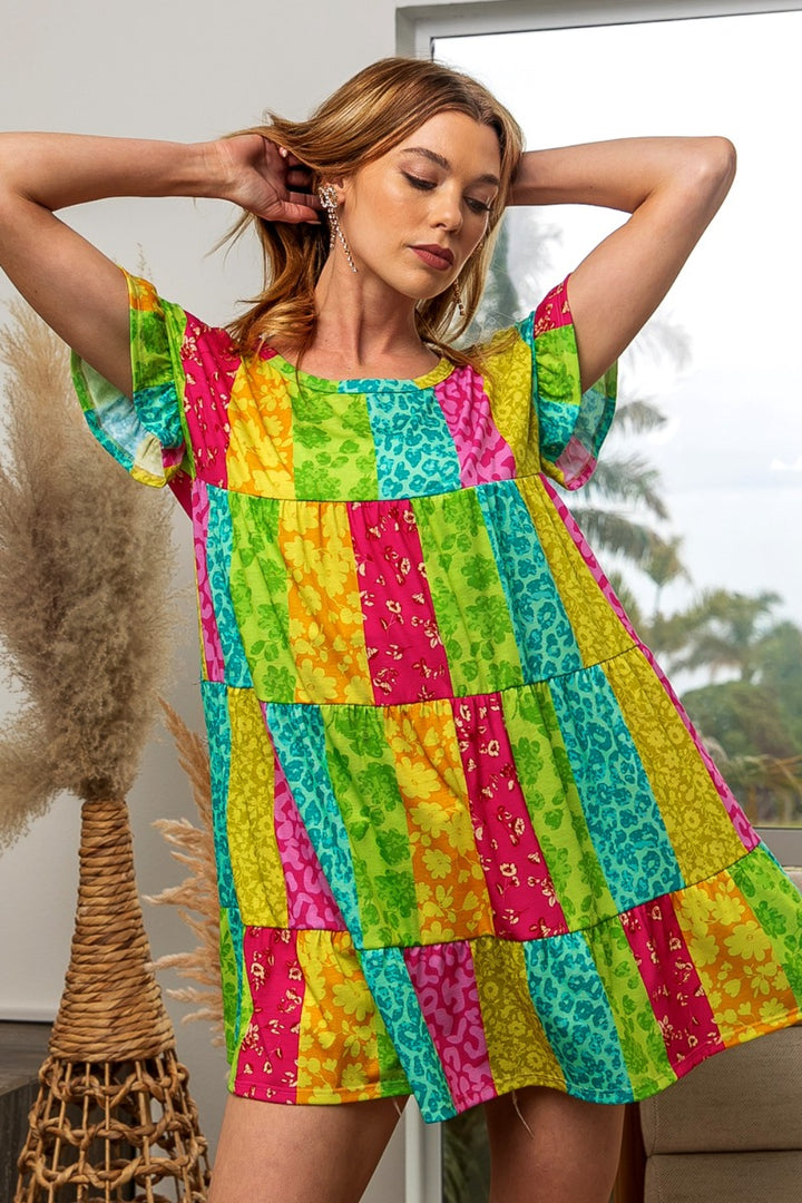 A woman in a BiBi Leopard Short Sleeve Tiered Dress, featuring colorful patchwork with floral and leopard patterns, stands indoors near a plant. One arm is raised behind her head near a window showing palm trees outside.