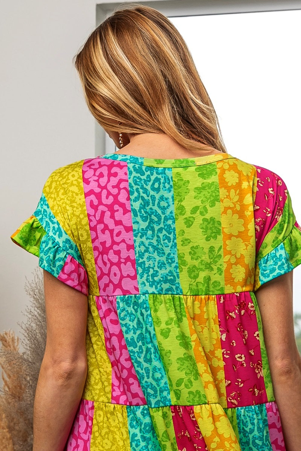 A woman in a BiBi Leopard Short Sleeve Tiered Dress, vibrant with pink, blue, yellow, and green vertical stripes among floral patterns, stands with her back to the camera. Her long hair is blonde.