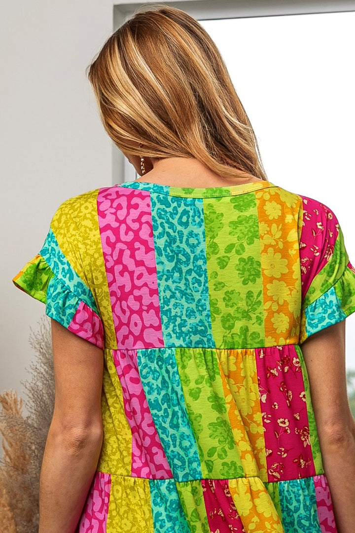 A woman in a BiBi Leopard Short Sleeve Tiered Dress, vibrant with pink, blue, yellow, and green vertical stripes among floral patterns, stands with her back to the camera. Her long hair is blonde.