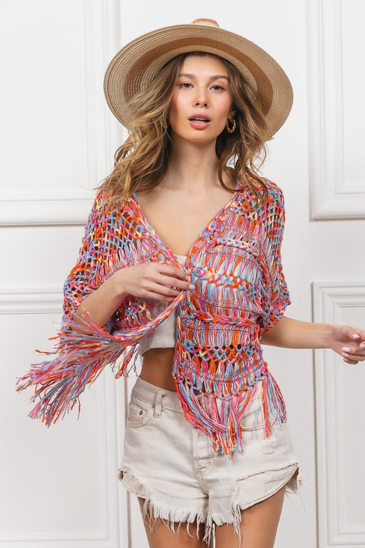 A woman in a BiBi Open Front Fringed Crop Knit Cardigan, wide-brimmed straw hat, and white shorts stands against a white paneled background. She has long wavy hair and gazes forward with a relaxed expression.