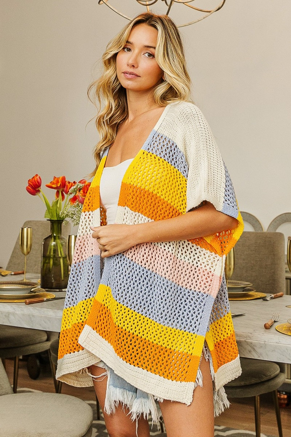 In a dining room adorned with a floral centerpiece and elegant table settings, a person wears the BiBi Openwork Striped Open Front Knit Cardigan over a white top and denim shorts. A brass chandelier hangs above, casting a warm glow over the scene.