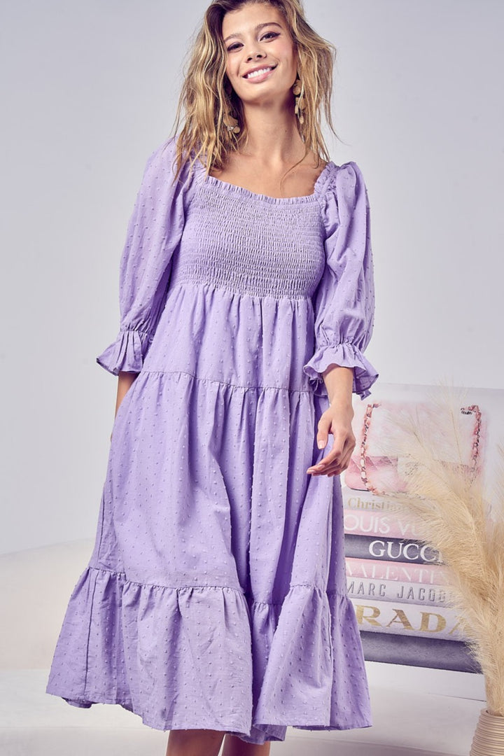 A woman smiles in the BiBi Swiss Dot Flounce Sleeve Smocked Tiered Midi Dress, standing in a chic room with beige and pink accents, a patterned pillow, and a wooden decor piece.