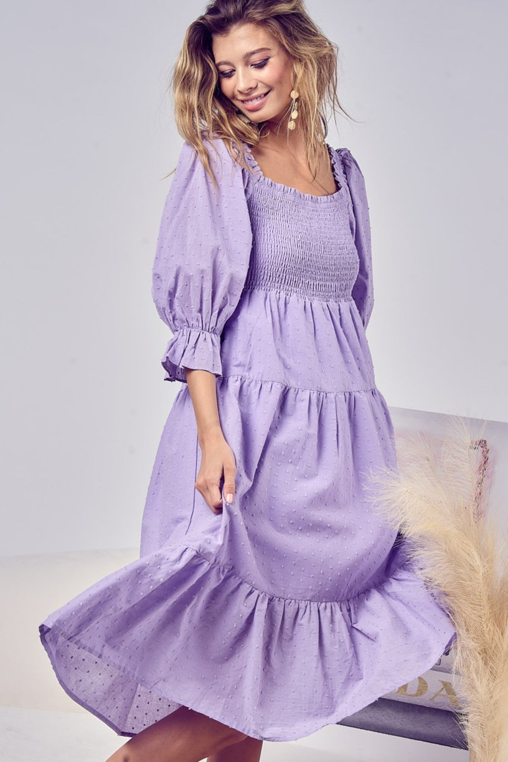 A woman indoors, with long hair and a subtle smile, holds the hem of her BiBi Swiss Dot Flounce Sleeve Smocked Tiered Midi Dress in lavender. The dress features a Swiss dot pattern and puff sleeves, while pampas grass decor is visible in the background.