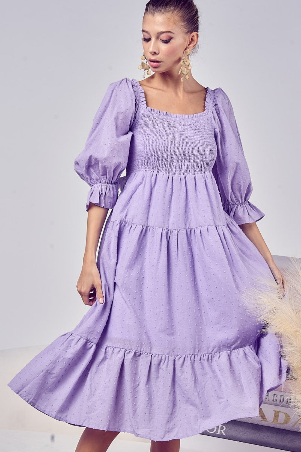 A woman models the BiBi Swiss Dot Flounce Sleeve Smocked Tiered Midi Dress in purple, glancing down while adorned with statement earrings. The neutral backdrop features decorative pampas grass.