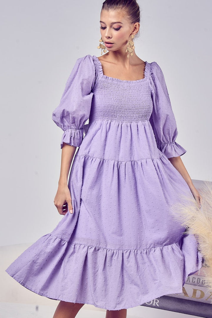 A woman models the BiBi Swiss Dot Flounce Sleeve Smocked Tiered Midi Dress in purple, glancing down while adorned with statement earrings. The neutral backdrop features decorative pampas grass.