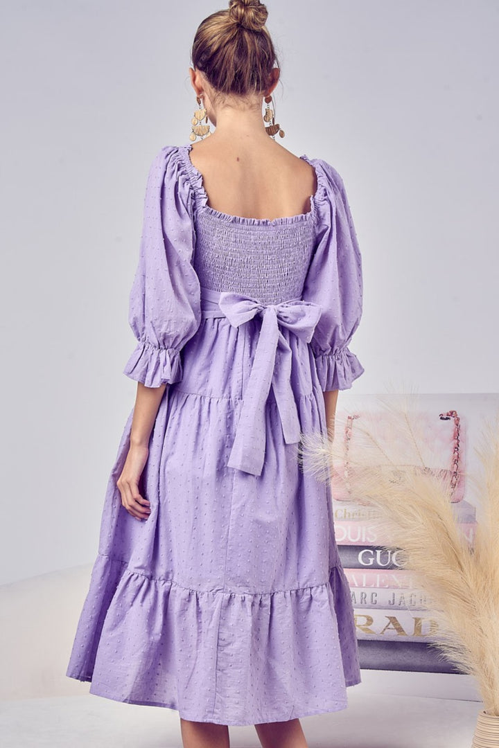 A woman wears the BiBi Swiss Dot Flounce Sleeve Smocked Tiered Midi Dress in lavender, featuring a smocked bodice, tiered skirt, and large back bow. The knee-length dress has flounce sleeves and Swiss dot texture. She stands in a softly lit room with fashion books nearby.
