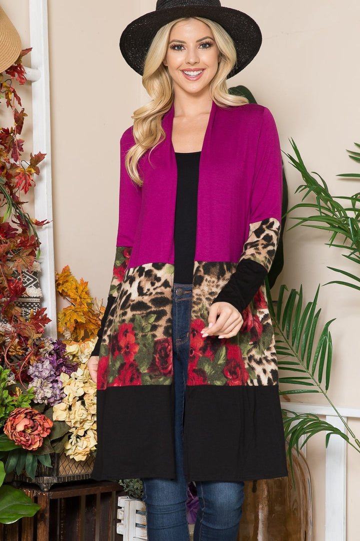 A woman wearing a wide-brimmed hat and the Celeste Full Size Color Block Open Front Cardigan, featuring magenta, leopard print, and floral accents, smiles at the camera. She is standing in a beautifully decorated space filled with colorful floral arrangements and greenery.