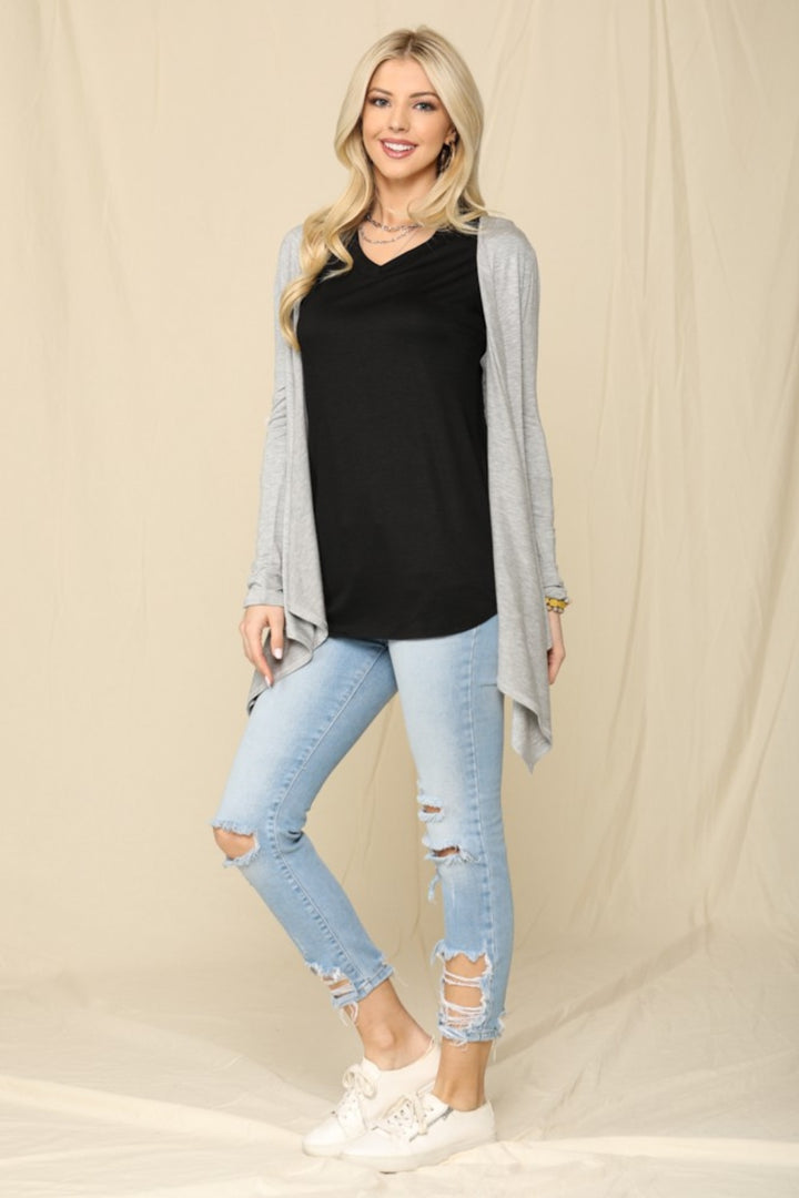 A woman in a black top, versatile light gray Celeste Full Size Open Front Knit Cardigan, and ripped light blue jeans poses against a beige curtain. She sports white sneakers and has long blonde hair.