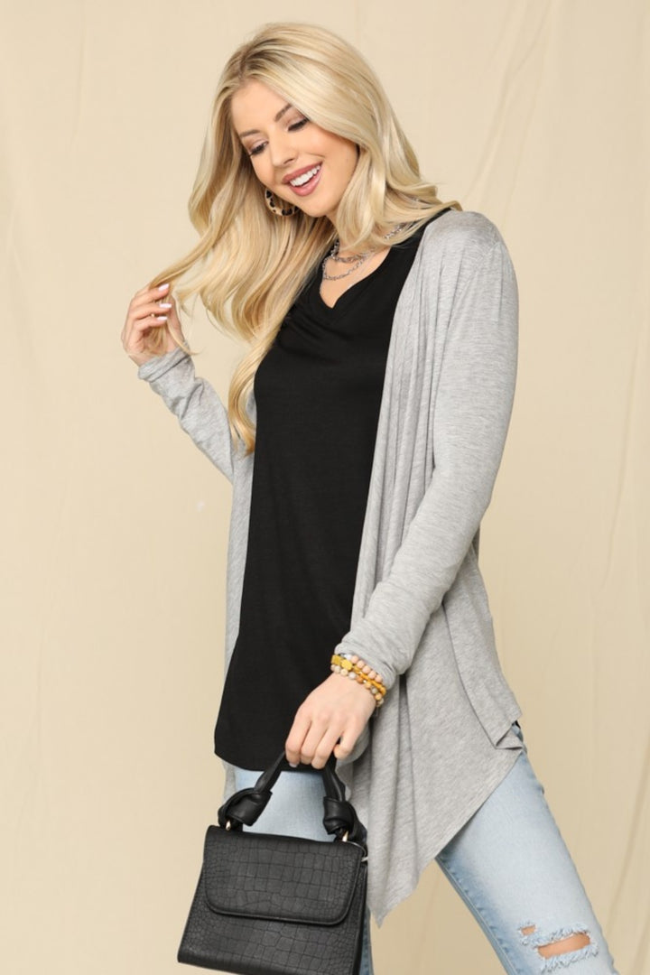A woman with long blonde hair smiles while holding a black handbag. She wears a black top, the Celeste Full Size Open Front Knit Cardigan, light blue jeans, and bracelets. The simple beige background emphasizes her versatile wardrobe staple.
