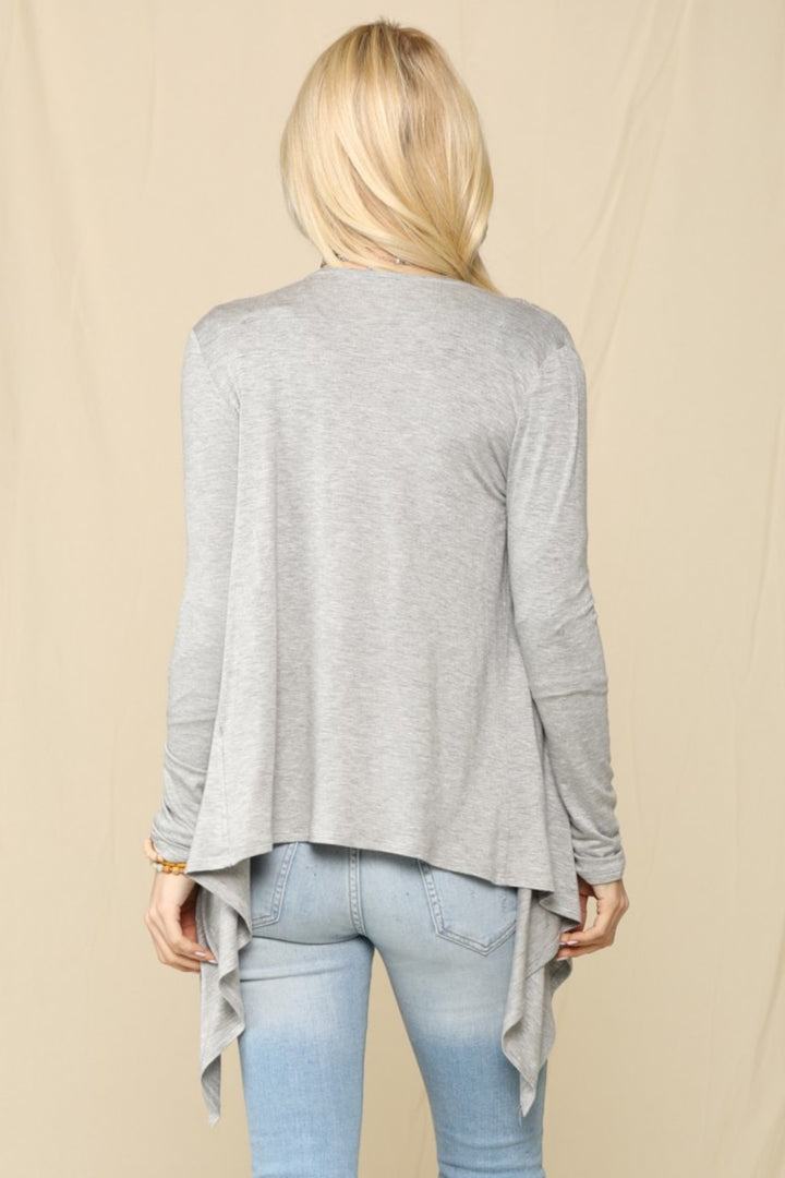 A person with long blonde hair, facing away, is wearing a Celeste Full Size Open Front Knit Cardigan in gray with an asymmetrical hem and light blue jeans. This versatile staple stands out against the plain beige background.