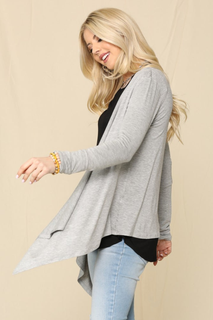 A blonde woman smiles while looking left, wearing the versatile Celeste Full Size Open Front Knit Cardigan in grey over a black shirt with blue jeans and bracelets, all set against a plain beige background.