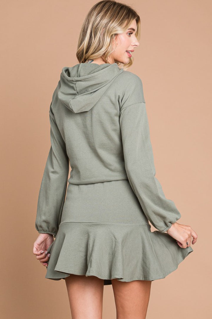 A woman models the Culture Code Full Size Drawstring Dropped Shoulder Hooded Mini Dress, featuring an olive green hue, long sleeves, and a flared hem. She faces slightly to the side, gently lifting the hem against a plain beige backdrop.