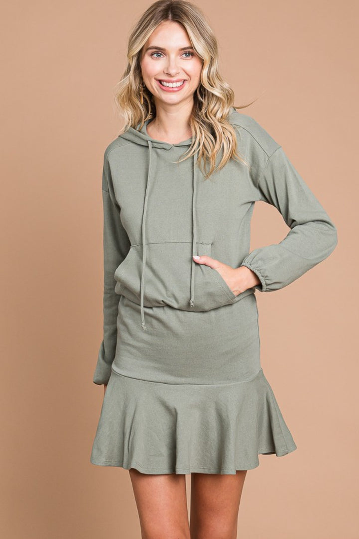 A woman in a Culture Code Full Size Drawstring Dropped Shoulder Hooded Mini Dress stands against a beige background. The light green mini dress has a front pocket and ruffled hem. With long, wavy blonde hair and one hand on her hip, she smiles, exuding casual elegance.