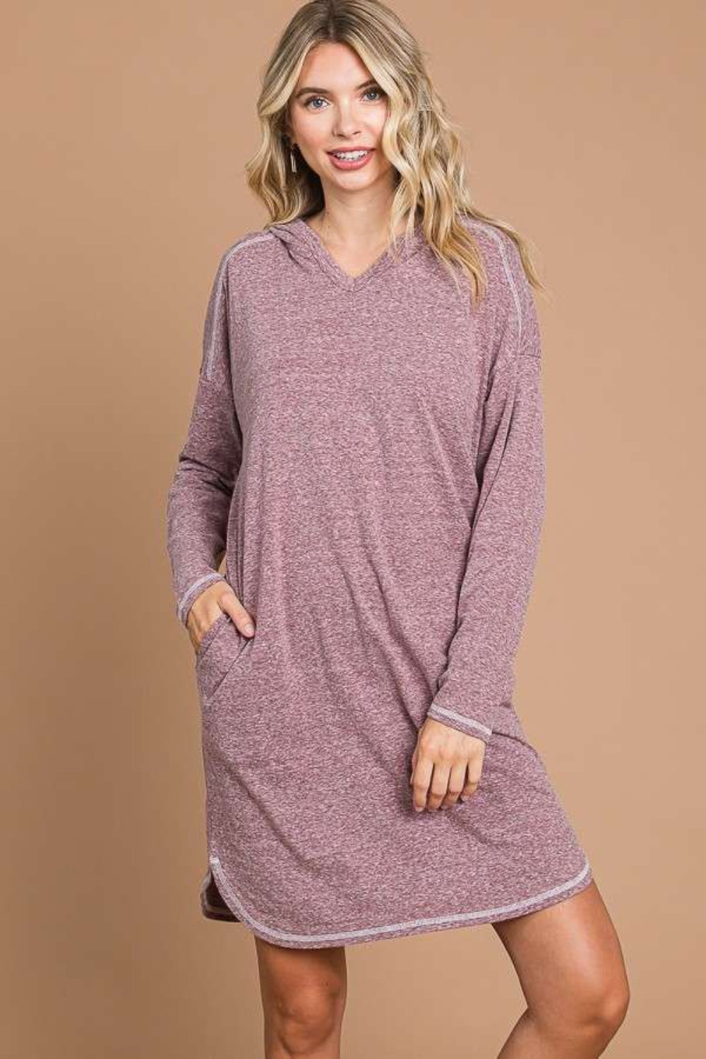 Against a plain brown background, an individual models the versatile Culture Code Full Size Hooded Long Sleeve Sweater Dress in cozy light mauve. With a hand casually tucked in their pocket and a smile directed at the camera, they effortlessly highlight this comfortable wardrobe essential.