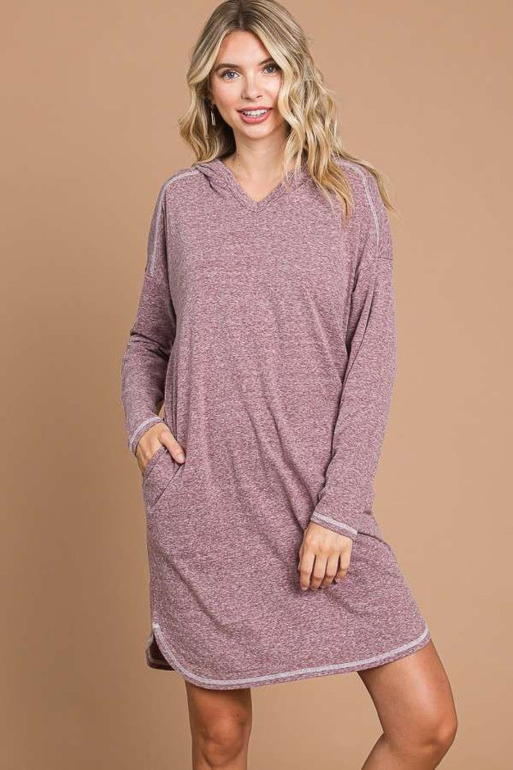 Against a plain brown background, an individual models the versatile Culture Code Full Size Hooded Long Sleeve Sweater Dress in cozy light mauve. With a hand casually tucked in their pocket and a smile directed at the camera, they effortlessly highlight this comfortable wardrobe essential.