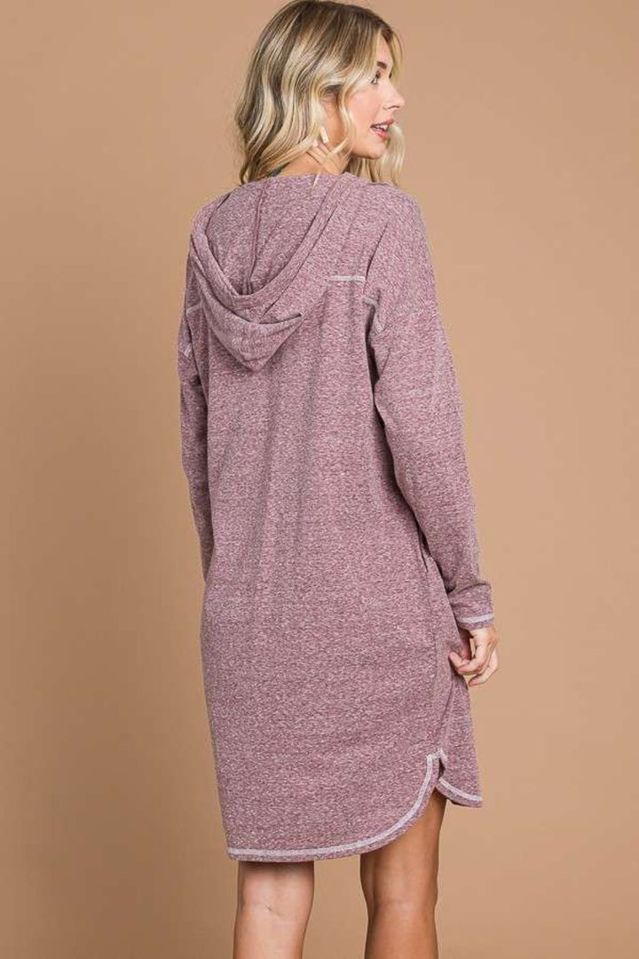 A person with long blonde hair is wearing the Culture Code Full Size Hooded Long Sleeve Sweater Dress in pink, crafted from cozy materials, standing against a beige background, shown from the back.