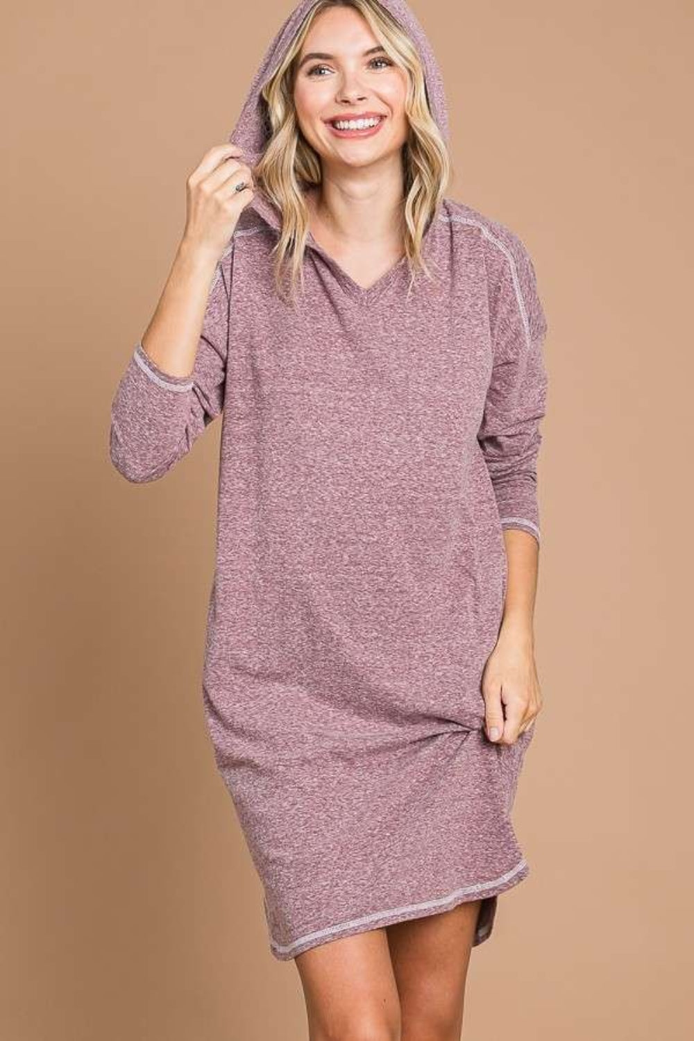 A person with blonde hair smiles while wearing the Culture Code Full Size Hooded Long Sleeve Sweater Dress in a mauve hue against a neutral background. They hold the hem of this cozy outfit with one hand and adjust the hood with the other, showcasing its versatility for any wardrobe.