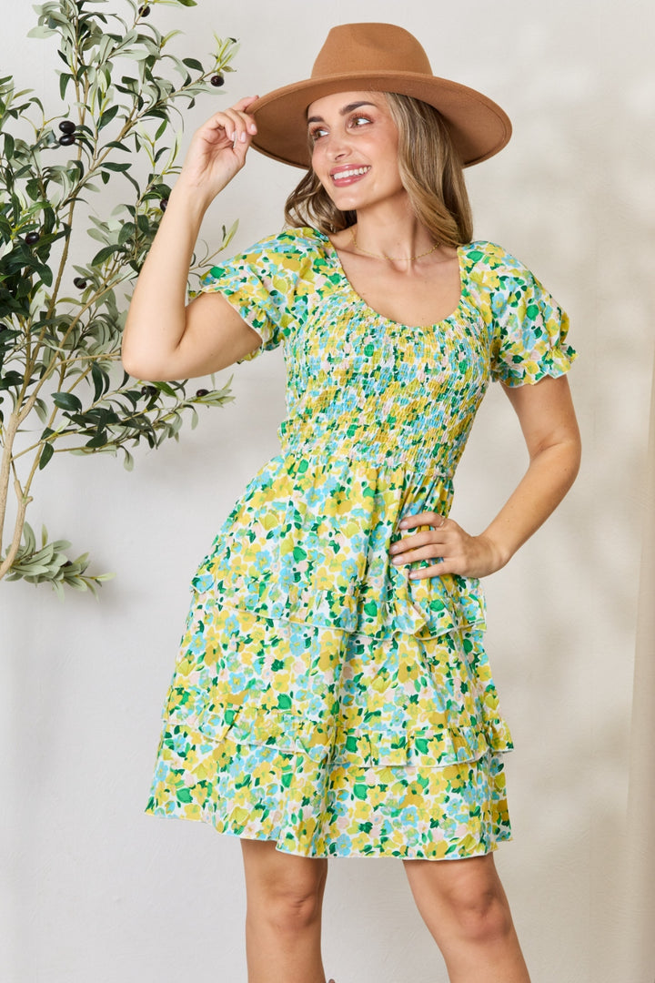 A woman smiles as she poses in the Devine Smocked Scoop Neck Flounce Sleeve Mini Dress, featuring a vibrant green and yellow floral pattern. One hand rests on her hip while the other adjusts her brown hat. She stands indoors against a light-colored wall, with a leafy plant beside her, highlighting the scoop neck design of her outfit.