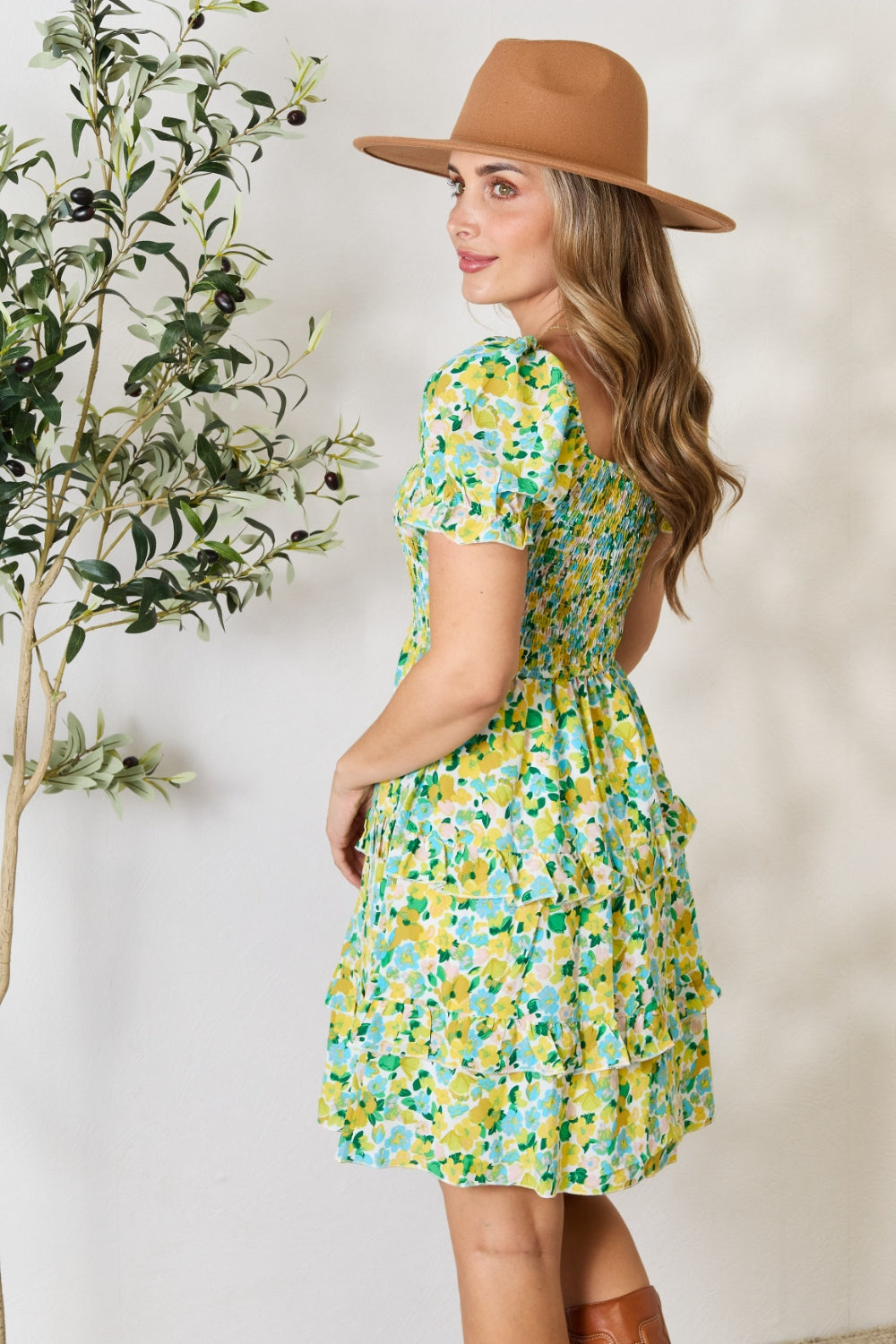 A person wearing the Devine Smocked Scoop Neck Flounce Sleeve Mini Dress and a brown hat stands next to a small olive tree. They have long hair and are looking over their shoulder with a serene expression.