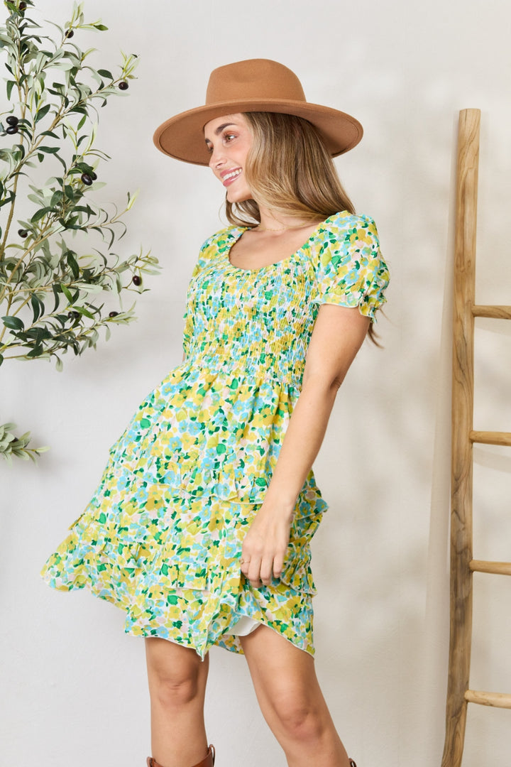A woman wearing the Devine Smocked Scoop Neck Flounce Sleeve Mini Dress smiles in her floral-patterned outfit. She pairs it with a brown hat and boots, standing beside a ladder and a leafy plant against a light-colored wall.