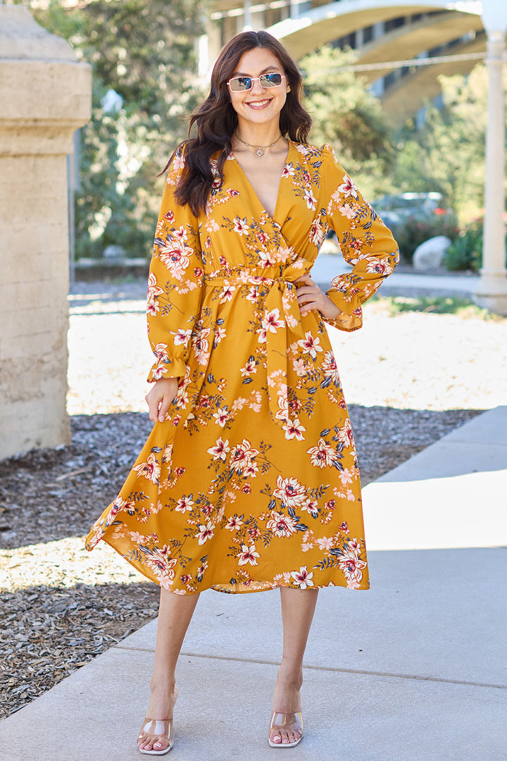 Wearing a Double Take Full Size Floral Tie Back Flounce Sleeve Dress, a woman in an inclusive size smiles under the sun on a path. Sporting sunglasses and open-toed heels, she poses with a hand on her hip. Trees and a stone structure adorn the backdrop. The easy-care dress is machine washable cold.