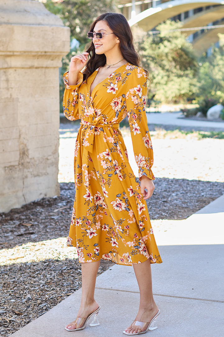 In a size-inclusive Double Take Floral Tie Back Flounce Sleeve Dress and clear heels, a woman stands outside smiling and looking sideways. She wears sunglasses and holds her hand near her chin, with a stone structure and greenery accentuating the beauty of her polyester outfit.