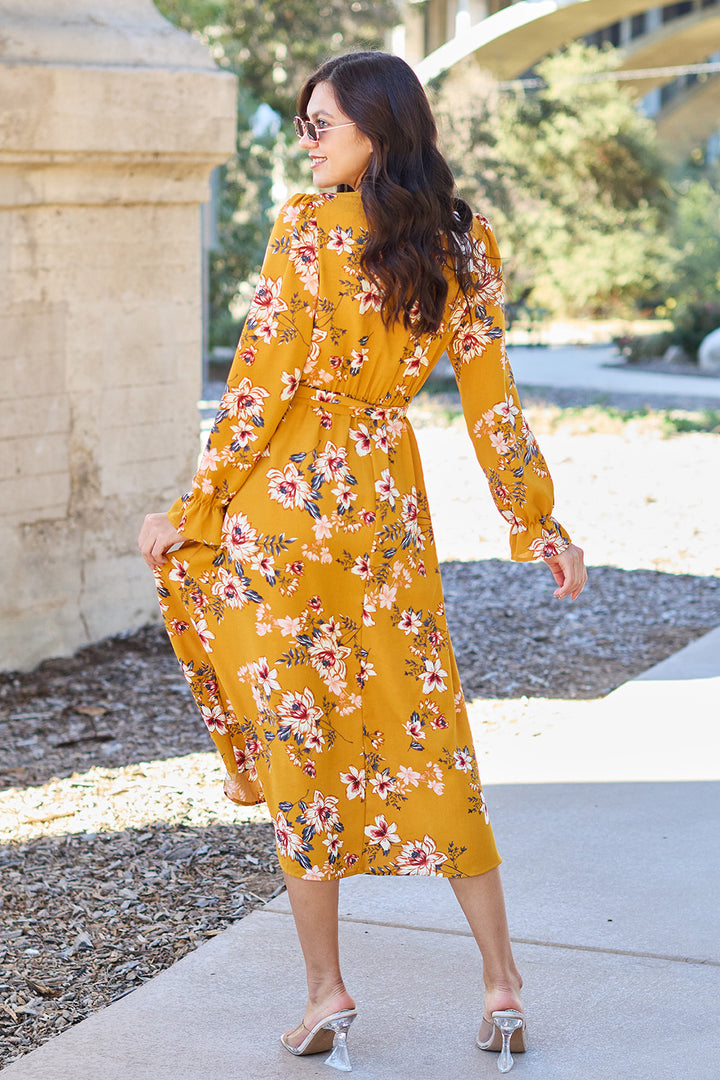 A woman with long wavy hair strolls outdoors on a sunny day, wearing sunglasses and the Double Take Full Size Floral Tie Back Flounce Sleeve Dress. She gently holds the dress at her side and wears transparent heels, smiling over her shoulder.