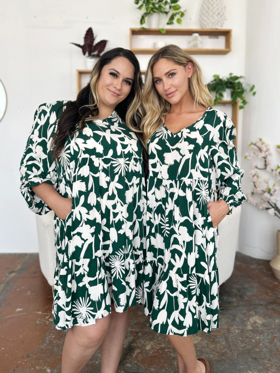 Two women stand side by side, smiling in matching Double Take Full Size Printed Ruffle Hem Dresses with a charming pocketed design. The cozy indoor setting is adorned with plants on shelves and a vase of flowers beside them, enhancing the appeal of their green and white floral rayon outfits.