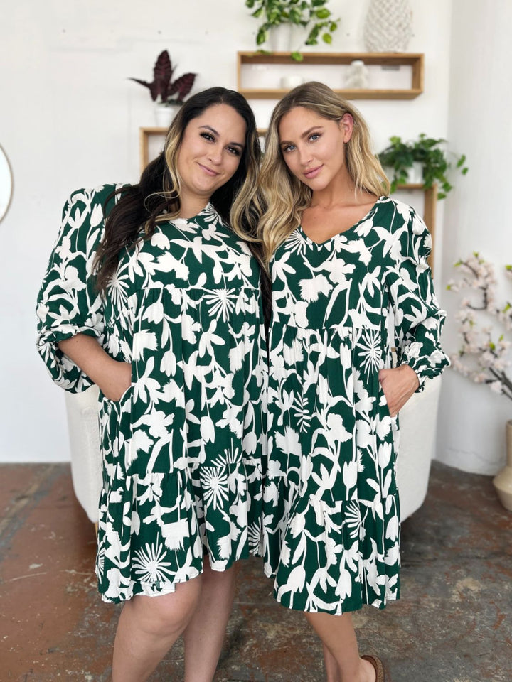 Two women stand side by side, smiling in matching Double Take Full Size Printed Ruffle Hem Dresses with a charming pocketed design. The cozy indoor setting is adorned with plants on shelves and a vase of flowers beside them, enhancing the appeal of their green and white floral rayon outfits.