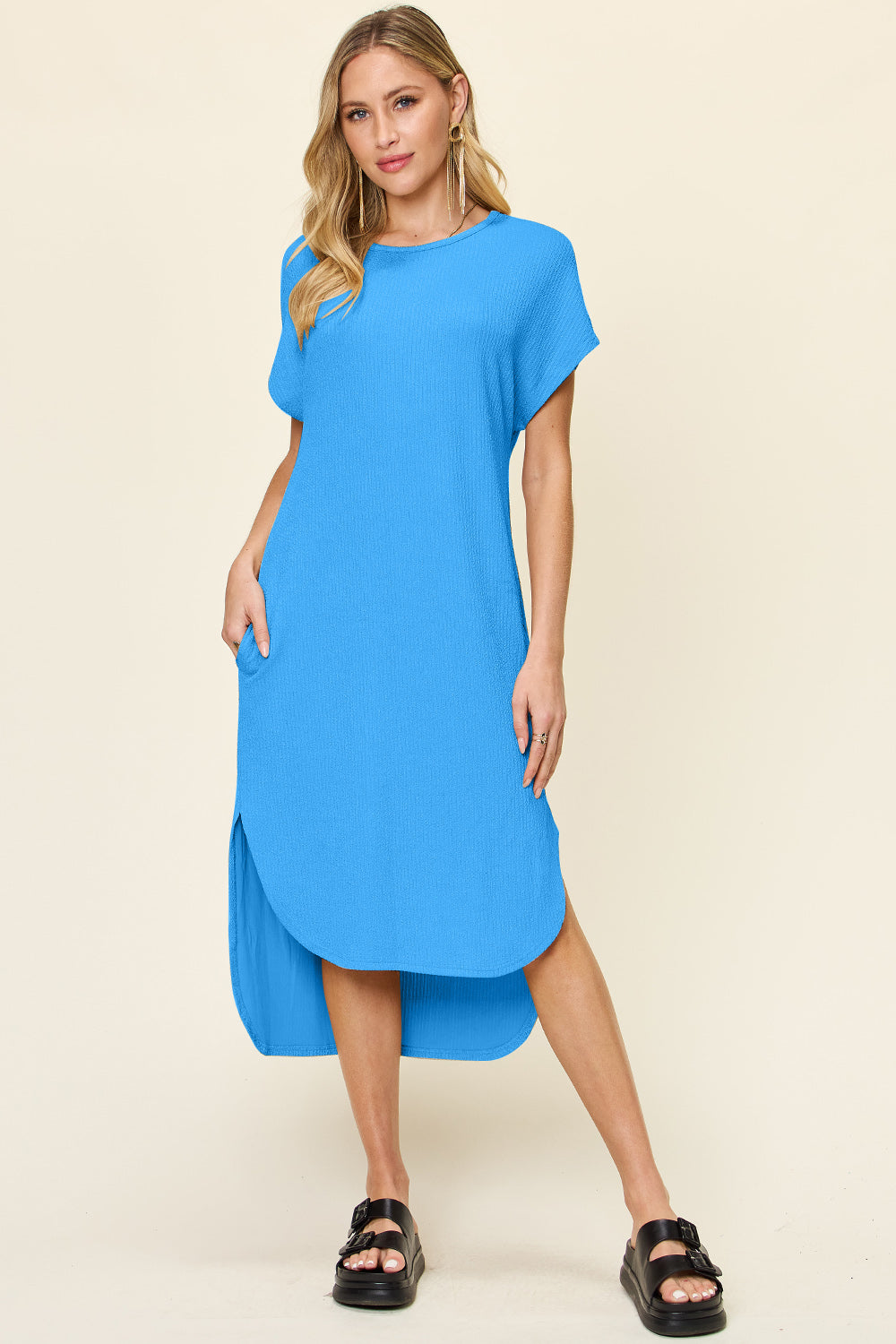 A woman with long blonde hair stands against a neutral background wearing hoop earrings, black slide sandals, and the Double Take Full Size Round Neck Short Sleeve Slit Dress in blue. Her right hand rests casually in her pocket.